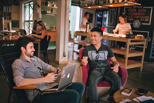 Two men socializing in a hostel, with people cooking in background.