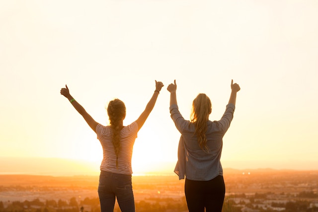 Two girls holding a thumbs up while watching a sunset. 